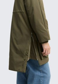 Giacca verde oliva con una finitura liscia e testurizzata. Presenta aperture laterali con zip e una vestibilità rilassata, abbinata a pantaloni in denim blu.