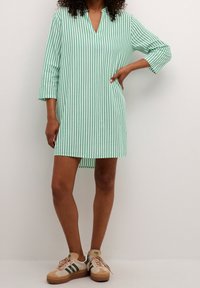 Sukienka-tunika w zielono-białe paski z dekoltem w kształcie litery V, rękawami do łokcia i krótszym przodem, zestawiona z beżowo-zielonymi sneakersami.