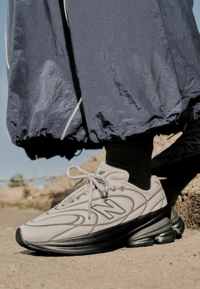 Baskets New Balance blanches et noires portées avec des chaussettes noires et un pantalon bleu ample, debout sur une surface en béton à l'extérieur.