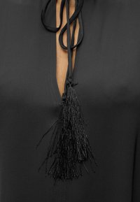 Μαύρο ύφασμα με βαθύ V ντεκολτέ που διαθέτει δύο μακριά κρόσσια κρεμασμένα από λεπτά κορδόνια δεμένα στο γιακά.