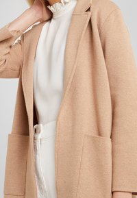 Beiger Strickcardigan mit offenem Vorderteil, langen Ärmeln und zwei Fronttaschen. Texturiertes Material mit einem weichen, warmen Aussehen.