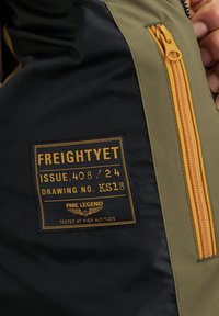Olive green and black jacket with orange zipper. Features a black label with gold text and winged emblem, indicating brand and specifications.