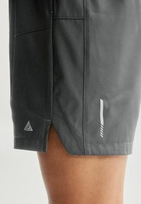 Grå træningsshorts med en glat tekstur, der har på siden slids, perforeret design og hvide logo detaljer nær kanten og på siden.