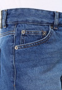 Sininen farkkuhousu klassisella viiden taskun muotoilulla, nappi- ja vetoketjusuljenta, erottuva oranssi ommel ja teksturoitu pinta.