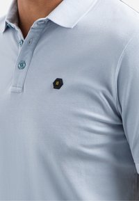 Lichtblauwe polo met een textuurstof, twee knopen en een zwart hexagonaal logo op de linkerkant van de borst. Contrasterende knopen als detail.