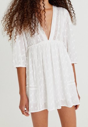 Robe blanche en coton avec un décolleté en V profond, manches courtes bouffantes, texture brodée, taille froncée et accents en dentelle sur les manches et l'ourlet.