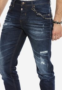 Dunkelblaue Jeans aus Denim mit einer taillierten Passform, die leichte Abnutzungsspuren, fransige Einsätze, kontrastierende gelbe Nähte und eine Kettenakzentuierung aufweisen.