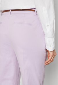 Personne portant un pantalon violet clair avec des poches passepoilées à l’arrière, une ceinture marron et une chemise blanche à manches longues avec poignets boutonnés.