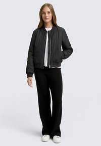 Sort quiltet bomberjakke med lynlås foran, tilpassede manchetter og talje, båret over en hvid top, kombineret med sorte bukser og hvide sneakers.