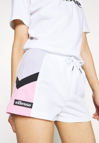 Shorts blancs avec une taille élastiquée et un cordon de serrage présentent des panneaux roses, noirs et gris. Un patch logo "ellesse" est visible sur la jambe gauche.