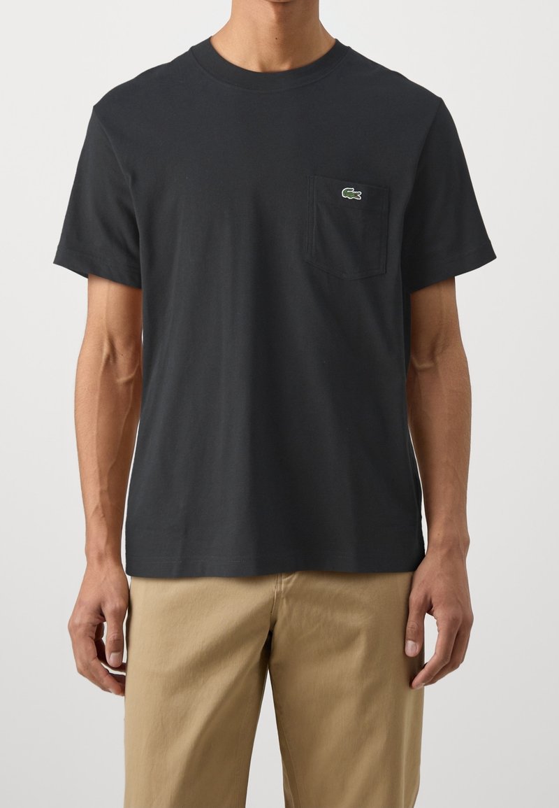 Black cotton t-shirt with short sleeves and a crew neckline. It features a left chest pocket and a green lacoste logo accent.