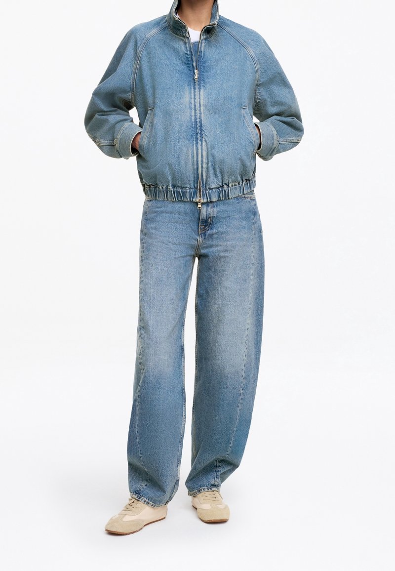 Personne portant une veste en jean bleu clair avec fermeture éclair, jean large assorti et baskets beiges, debout les mains dans les poches de la veste.