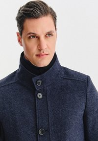 Granatowy płaszcz wełniany w mieszance z wysokim kołnierzem, dwoma dużymi guzikami z przodu i teksturowanym materiałem. Projekt jest dopasowany i strukturalny.