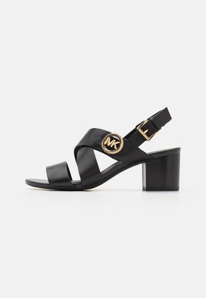 Sort læder åben tå sandal med blokhæl, krydsede stropper, guldfarvet spænde og en rund MK-logo detalje på siden af stroppen.