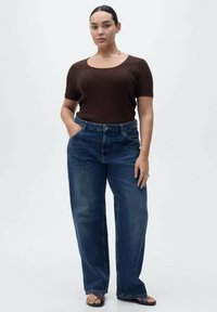 Top corto a maniche corte marrone a costine abbinato a jeans a gamba larga blu a vita alta. Modella mostrata con le mani in tasca, sandali flat e gioielli minimal.