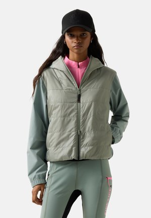 Woman wearing black cap, light green zip-up jacket with hood, pink top underneath, and matching light green pants with pink zipper detail.