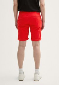 Pantalones cortos de algodón rojo con puños doblados, detalle de bolsillo trasero, combinados con zapatillas blancas y calcetines hasta el tobillo, vistos desde la parte trasera.