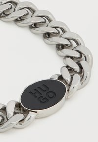 Ασημένιο βραχιόλι αλυσίδα με στρογγυλεμένους συνδέσμους και μαύρη οβάλ ετικέτα με ανάγλυφη τη λέξη «Hugo». Λείο υλικό μετάλλου, γυαλιστερό φινίρισμα.