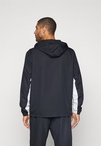 Čierna bunda s kapucňou s bielymi bočnými panelmi, vybavená kapucňou so sťahovacou šnúrkou, elastickými manžetami a hladkou, ľahkou textúrou látky.