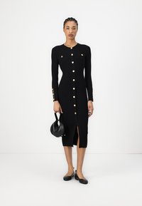 Robe en tricot côtelé noire à manches longues, avec des boutons dorés sur le devant, une fente à l'ourlet et deux poches avant. Accessoire : petit sac noir.