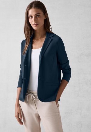 Blazer marine avec une fermeture à un bouton, revers crantés, deux poches avant et un design ajusté, porté sur un haut blanc et un pantalon clair.