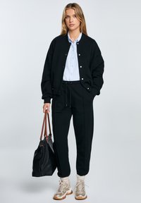 Černá oversized bomber bunda, světle modrá košile na knoflíky, černé kalhoty na šnůrku, zlaté tenisky a černá kožená taška na rameno.