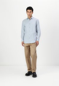 Camicia a righe azzurra e bianca con colletto a punta, abbinata a pantaloni beige e scarpe nere su uno sfondo neutro.