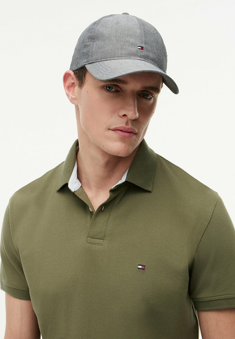 Jeune homme portant une casquette de baseball grise et un polo vert olive avec un petit logo rouge, blanc et bleu sur la poitrine.