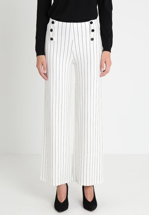 Vrouw draagt witte broek met wijde pijpen met zwarte verticale strepen en vier zwarte knopen aan elke zijde ter hoogte van de taille, gecombineerd met zwarte hakken.