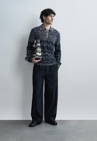 Mand stående op ad væggen iført mønstret marineblå sweater, brede sorte bukser, sorte sko, holder en sort-hvid zigzag mønstret taske.