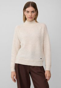 Kobieta nosząca sweter w kremowym kolorze z fakturowanym splotem, z prążkowanymi mankietami i kołnierzem, w połączeniu z luźnymi brązowymi spodniami, stojąca na jednolitym tle.