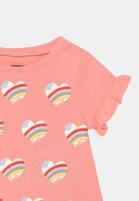 T-shirt de algodão rosa claro com um padrão repetido de corações com riscas em arco-íris e um detalhe de nuvem azul. Mangas curtas com volantes.