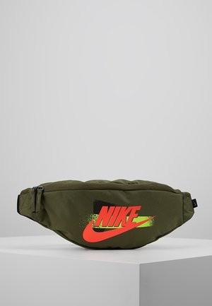 Sac banane Nike vert olive avec un logo rouge vif et jaune, fermeture éclair noire et sangle réglable, présenté sur un socle blanc.