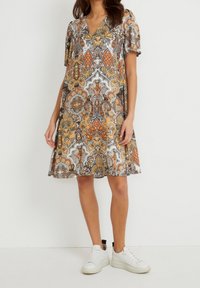 Kortsleeve, v-ringad klänning med ett flerfärgat paisleymönster i orange, brunt och vitt, med en utsvängd nederkant. Bärs med vita sneakers.