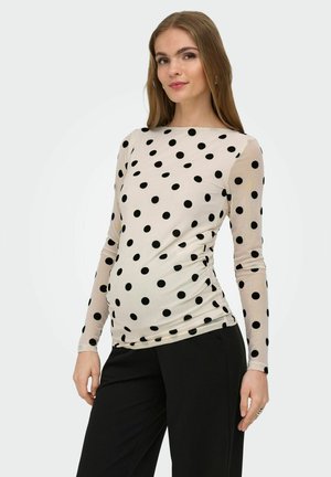 Gravid kvinde iført en beige langærmet top med sorte polkaprikker og sorte bukser, stående med den ene hånd afslappet.