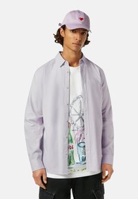 Chemise à rayures violettes claires sur un t-shirt graphique blanc avec des illustrations colorées, associée à une casquette lavande arborant un cœur rouge.