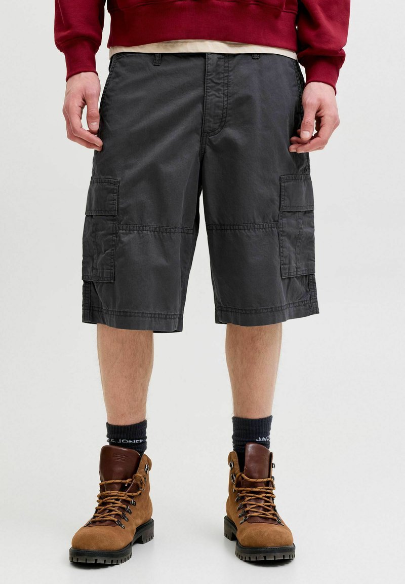 Personne portant un short cargo noir, des chaussettes noires, des bottes de randonnée marron et un sweat-shirt rouge, debout avec les mains détendues le long du corps.