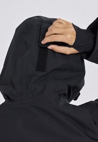 Giacca nera impermeabile con cappuccio e chiusura in Velcro. Il tessuto appare liscio con una finitura opaca e cuciture minime visibili.