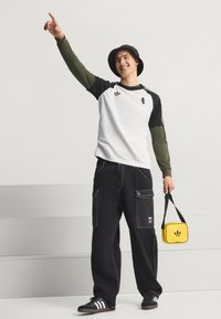 Μαύρο παντελόνι καρό με λευκά ραφές, λευκό και μαύρο μακρυμάνικο μπλουζάκι και κίτρινη τσάντα. Το άτομο φοράει μαύρο καπέλο τύπου bucket και αθλητικά παπούτσια.