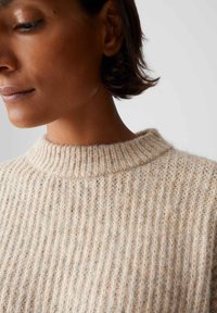Donna con capelli corti e scuri che indossa un maglione a coste beige, mostrata dal collo alla parte superiore del petto su uno sfondo semplice.
