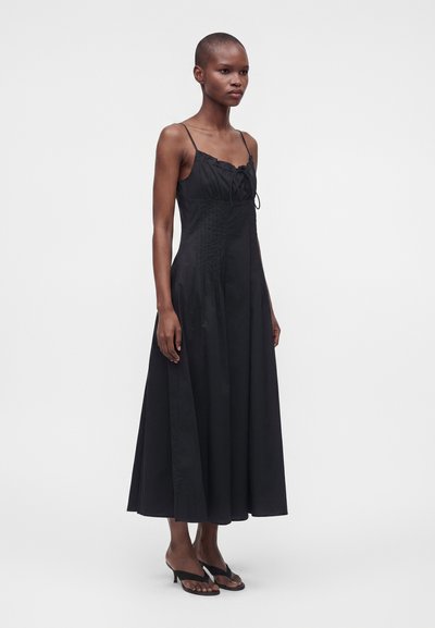 Femme aux cheveux courts portant une robe longue noire sans manches avec de fines bretelles et des sandales à talons noires à bout ouvert, debout sur un fond uni.