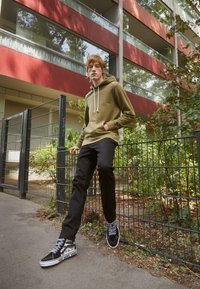 Felpa verde oliva con lacci bianchi, jeans neri e sneakers Vans nere con dettagli bianchi, in piedi di fronte a una recinzione e alla vegetazione.