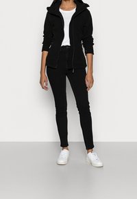 Felpa nera con zip in materiale morbido, abbinata a jeans neri attillati e sneaker bianchi. L'outfit presenta una semplice maglietta bianca sotto.