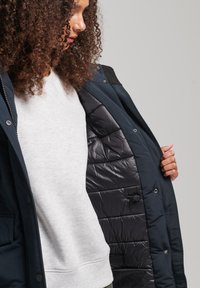 Marineblauwe geïsoleerde jas met ritsluiting, knoopdetails en een glanzend zwarte voering. Model draagt een lichtgrijze sweatshirt eronder.