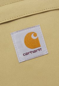 Parche del logo de Carhartt con emblema naranja y texto marrón sobre un fondo de tela beige y duradera, cosido en una prenda o bolsa.