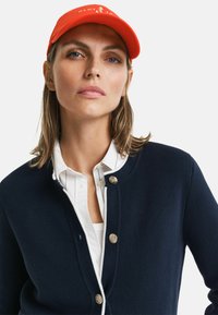 Marineblauer Cardigan mit strukturierter Oberfläche und silbernen Knöpfen, über einem weißen gestreiften Hemd getragen und mit einer leuchtend orangefarbenen Mütze ergänzt.