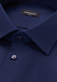 Camicia da uomo blu navy con tessuto liscio, colletto a punta e chiusura con un bottone caratterizzato da un design a quattro fori. Presente etichetta Seidensticker.