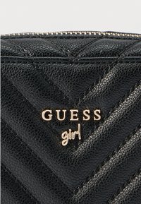 Musta tikattu lompakko, jossa on teksturoitu keinonahka, kultainen "GUESS"-logo ja "girl"-koriste. Vetoketjusuljenta koristeellisilla metalliyksityiskohdilla.