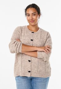 Beżowy sweter z dzianiny o teksturowanym wzorze, okrągłym dekolcie i czarnych guzikach w kształcie kokardy, w połączeniu z jasnoniebieskimi dżinsami. Miękki i przytulny materiał.