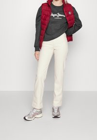Gilet imbottito rosso sopra una felpa scura con testo bianco, abbinato a pantaloni larghi color crema e scarpe sportive argento con dettagli.